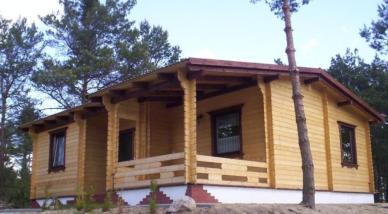 Gotowy, parterowy dom z bali o jasnej elewacji, z zadaszonym tarasem wspartym na drewnianych kolumnach, widoczny na tle sosnowego lasu.